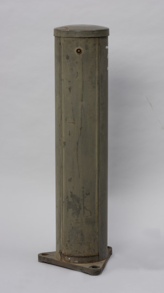 Systemowe Urządzenie Sygnalizacyjne - Ujęcie z tyłu; Urządzenie sygnalizacyjne będące fragmentem systemu ochrony granic PRL. Metalowy słup, w którym zamontowano czujniki na podczerwień. Obudowa o kształcie walca zamontowana na trójkątnej podstawie.