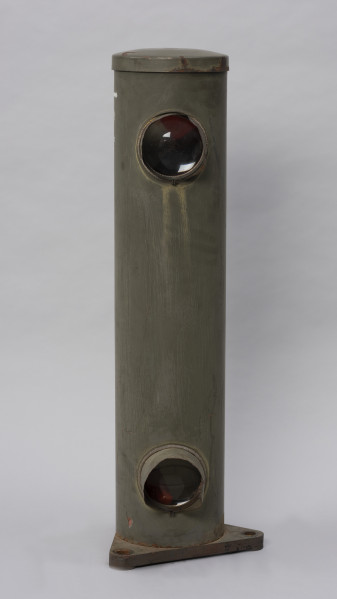 Systemowe Urządzenie Sygnalizacyjne - Ujęcie z przodu; Urządzenie sygnalizacyjne będące fragmentem systemu ochrony granic PRL. Składa się z dwóch metalowych słupów, w których zamontowano czujniki na podczerwień. Każde z urządzeń znajduje się w obudowie o kształcie walca zamontowanej na trójkątnej podstawie. Zaopatrzone jest w dwie soczewki umieszczone z przodu jedna nad drugą.