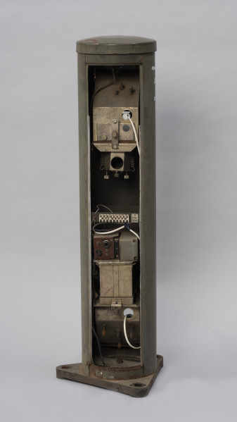 Systemowe Urządzenie Sygnalizacyjne - Ujęcie z tyłu; Urządzenie sygnalizacyjne będące fragmentem systemu ochrony granic PRL. Metalowy słup, w którym zamontowano czujniki na podczerwień. Obudowa o kształcie walca zamontowana na trójkątnej podstawie. Zaopatrzone jest w dwie soczewki umieszczone jedna nad drugą.