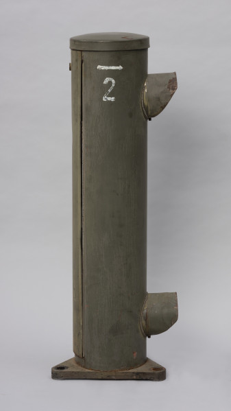 Systemowe Urządzenie Sygnalizacyjne - Ujęcie z lewej strony; Urządzenie sygnalizacyjne będące fragmentem systemu ochrony granic PRL. Metalowy słup, w którym zamontowano czujniki na podczerwień. Obudowa o kształcie walca zamontowana na trójkątnej podstawie. Zaopatrzone jest w dwie soczewki umieszczone jedna nad drugą z prawej strony. w górnej części malowany symbol strzałki skierowanej w prawą stronę a pod nią cyfra 2.