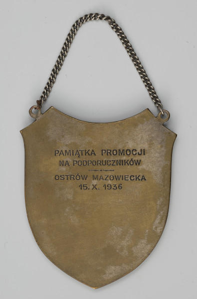 ryngraf z Matką Boską Ostrobramską - ujęcie z tyłu; Ryngraf w formie tarczy. Na odwrocie, w górnej części wyryty jest napis okolicznościowy. W górnej części, na ukośnych bokach tarczy dwa dolutowane oczka umożliwiające zawieszenie ryngrafu na łańcuszku. Łańcuszek wykonano z innego metalu, a jedno z ogniw, łączące łańcuszek z prawym oczkiem ryngrafu, dosztukowano z miedzianego drutu.