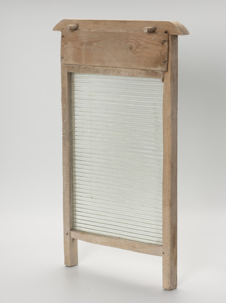 Tara do prania - ujęcie ze skosu od tyłu; Tara do prania złożona z prostokątnej, szklanej tafli osadzonej w drewnianej ramie. Szklana część jednostronnie karbowana. Oprawa z dwóch pionowych listew tworzących w dolnej części nóżki, dwóch poprzecznych listew stanowiących oparcie dla tafli oraz górnej profilowanej z wystającymi końcówkami stanowiącymi uchwyt. Od tyłu, pomiędzy listwą uchwytową, a górną poprzeczką przybita prostokątna, cienka deska. Powyżej w listwie uchwytowej wkręcone dwa drewniane kołki umożliwiające zawieszenie tary na brzegach balii.