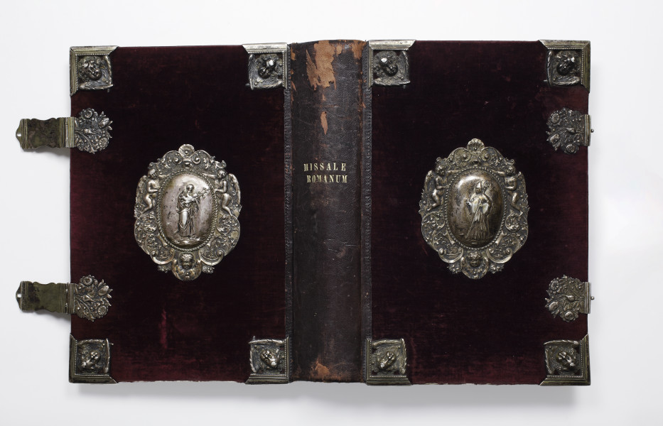 Fotografia kolorowa. Książka rozłożona tak, że w ujęciu na wprost widoczne są jej okładki, grzbiet i klamry do spięcia oprawy. Grzbiet skórzany, ciemnobordowy, z jaśniejszymi przetarciami. Mniej więcej pośrodku grzbietu wytłoczony złotymi literami, w dwóch rzędach, tytuł księgi MISSALE ROMANUM. Okładka obciągnięta aksamitem w kolorze ciemno bordowym. Zdobiona srebrnymi aplikacjami z wypukłymi wzorami. Pośrodku obu okładek owalny, srebrny medalion. W medalionach przedstawienia świętych. Na okładce tylnej, widocznej po lewej, postać mężczyzny w długiej szacie z dzieckiem na ręku. Na okładce przedniej, widocznej po prawej, postać mężczyzny w stroju biskupim z pastorałem w ręce i w szpiczastym nakryciu głowy. W otoku medalionów o nieregularnym kształcie motywy roślinne i dwa anioły. Na rogach okładek srebrne narożniki z uskrzydlonymi główkami aniołów. Z lewej strony, przy dłuższej krawędzi okładki, dwie srebrne klamry przytwierdzone okuciami zdobionymi motywami bukietów kwiatów. Na okładce z prawej strony identyczne okucia z zapięciem klamer.
