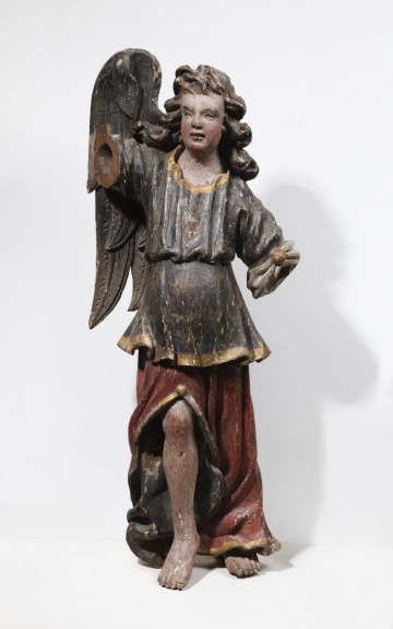 Fotografia kolorowa. Figura ołtarzowa, przyścienna, a więc płasko ścięta z tyłu, z pomalowanego w różnych kolorach drewna. Przedstawia stojącego anioła z jednym, prawym skrzydłem. Głowa uniesiona lekko ku górze i zwrócona w lewo. Prawe ramię uniesione ku górze, lewa ręka opuszczona w dół, odsunięta od ciała. Twarz pełna, o szerokim czole i policzkach, okolona bujnymi, długimi, przestrzennie rzeźbionymi falami włosów, opadających na ramiona. Nos krótki i szeroki, wydatne usta. Anioł odziany jest w brunatno czerwoną, długą szatę spodnią ze złotym oblamowaniem, rozciętą z przodu u dołu tak, że ukazuje ona prawą nogę do kolana. Na wierzch nałożona jest szara, przepasana tunika ze złotą lamówką o długich, luźnych rękawach upiętych guzami przy łokciach. Brak obu rąk poniżej łokci.