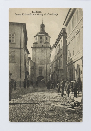 ML/H/2242 Na brukowanej ulicy i chodnikach grupy pieszych. Ulica w perspektywie, po obu stronach dwupiętrowe kamienice. Na zakończeniu ulicy Brama Krakowska. Brama otynkowana z żelazna galerią dla straży dookoła górnej kondygnacji. W górnej części podpis. Na odwrociu w górnej częsci napis