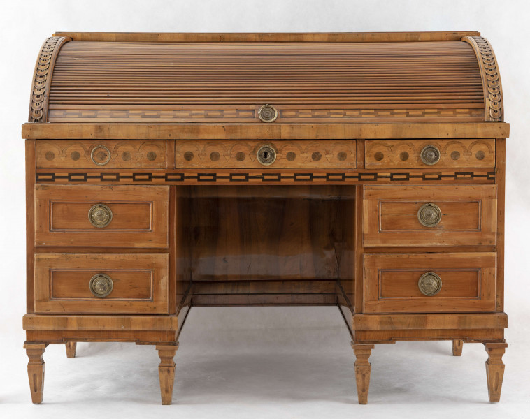 Biurko przyścienne na sześciu nóżkach graniastych zbieżnych. W części dolnej prostokątny prześwit zamknięty tylną ścianą. Po jego bokach po dwie szuflady, powyżej jeszcze trzy. Część górna ćwierćcylindryczna zamykana żaluzją chowaną pod górna płytą powyżej pięciu szufladek, boki zdobione plastycznym ornamentem cekinowym. Lico pokryte fornirem z intarsją ramkową i wzorem arkadkowym. Blat wyłożony skórą cielęcą. Uchwyty szuflad zewnętrznych i żaluzji to nałożone okucia okrągłe z tarczą, szufladki w części górnej mają uchwyty okrągłe z gładką główką.  