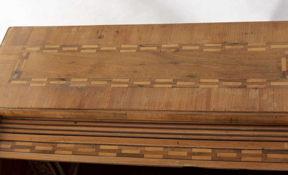 Biurko przyścienne na sześciu nóżkach graniastych zbieżnych. W części dolnej prostokątny prześwit zamknięty tylną ścianą. Po jego bokach po dwie szuflady, powyżej jeszcze trzy. Część górna ćwierćcylindryczna zamykana żaluzją chowaną pod górna płytą powyżej pięciu szufladek, boki zdobione plastycznym ornamentem cekinowym. Lico pokryte fornirem z intarsją ramkową i wzorem arkadkowym. Blat wyłożony skórą cielęcą. Uchwyty szuflad zewnętrznych i żaluzji to nałożone okucia okrągłe z tarczą, szufladki w części górnej mają uchwyty okrągłe z gładką główką.  