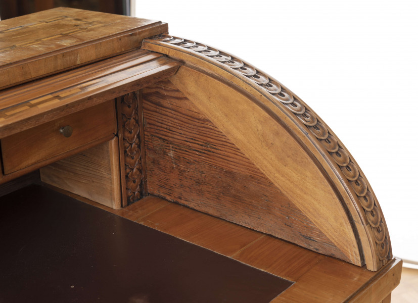 Biurko przyścienne na sześciu nóżkach graniastych zbieżnych. W części dolnej prostokątny prześwit zamknięty tylną ścianą. Po jego bokach po dwie szuflady, powyżej jeszcze trzy. Część górna ćwierćcylindryczna zamykana żaluzją chowaną pod górna płytą powyżej pięciu szufladek, boki zdobione plastycznym ornamentem cekinowym. Lico pokryte fornirem z intarsją ramkową i wzorem arkadkowym. Blat wyłożony skórą cielęcą. Uchwyty szuflad zewnętrznych i żaluzji to nałożone okucia okrągłe z tarczą, szufladki w części górnej mają uchwyty okrągłe z gładką główką.  