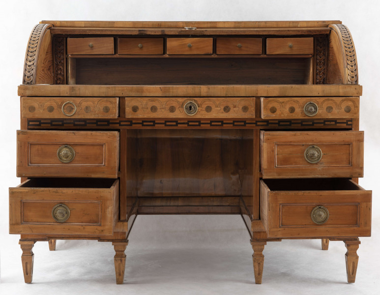 Biurko przyścienne na sześciu nóżkach graniastych zbieżnych. W części dolnej prostokątny prześwit zamknięty tylną ścianą. Po jego bokach po dwie szuflady, powyżej jeszcze trzy. Część górna ćwierćcylindryczna zamykana żaluzją chowaną pod górna płytą powyżej pięciu szufladek, boki zdobione plastycznym ornamentem cekinowym. Lico pokryte fornirem z intarsją ramkową i wzorem arkadkowym. Blat wyłożony skórą cielęcą. Uchwyty szuflad zewnętrznych i żaluzji to nałożone okucia okrągłe z tarczą, szufladki w części górnej mają uchwyty okrągłe z gładką główką.  