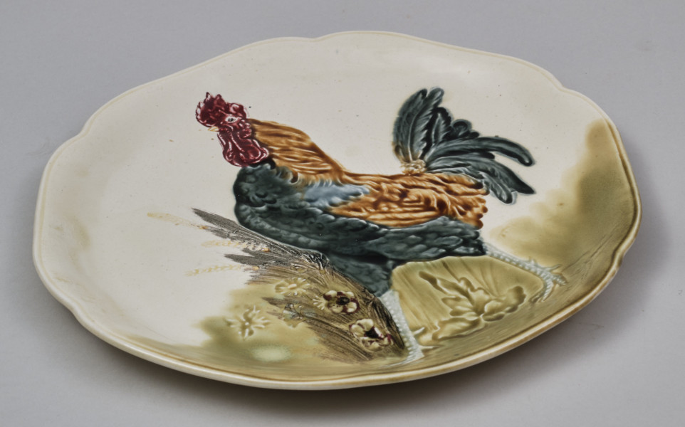 Talerz fajansowy płaski z łagodnie podniesionym brzegiem.
Głównym, centralnym motywem dekoracyjnym jest kogut.
