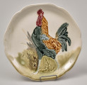Talerz fajansowy płaski z łagodnie podniesionym brzegiem.
Głównym, centralnym motywem dekoracyjnym jest kogut.