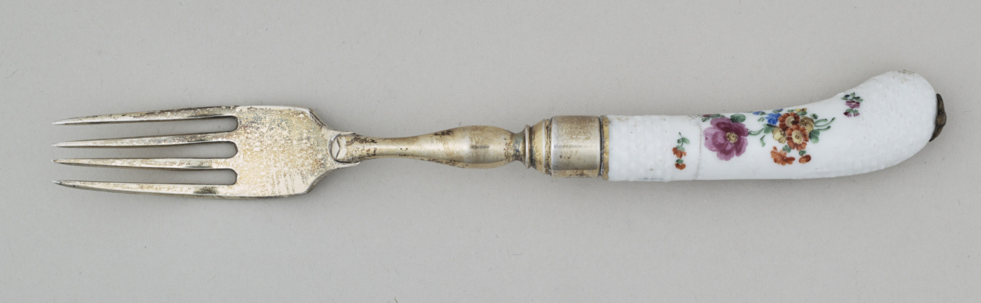 Widelec śniadaniowy, czterozębny. Zęby wygięte, zwężające się przy zakończeniu. Trzonek z nakładką ze srebra złoconego z białej porcelany