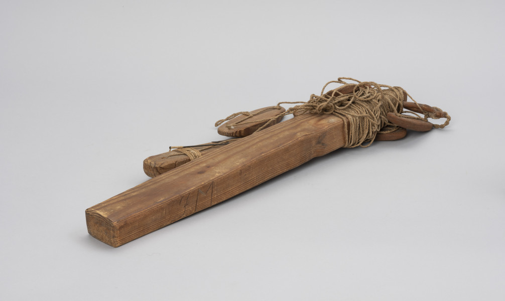 takla łososiowa z merkiem - Ujęcie ze skosu z prawej; Jednohaczykowa węda morska dryfująca służąca do połowu pojedynczych łososi znakowana merkiem FM.
Prezentowane narzędzie składa się z dużego, drewnianego pływaka (koby)   i sznura  oraz 7 pływaków - 2 wykonane z drewna  i 5 z kory. Koba  zakończona jest haczykiem z przynętą w postaci żywej rybki, której brak, obciążona jest stożkowatym ciężarkiem. Pływak - (koba) z drewna jest znakowany merkiem (znakiem własności) - inicjał FM ( być może są to inicjały darczyńcy).