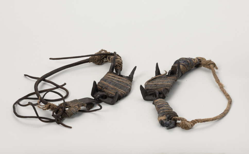 raki do chodzenia po lodzie - Ujęcie z przodu; Czteroramienne raki położone na widocznym ujęciu do góry nogami, wykonane techniką kowalską o ramionach na narożach hakowato odgiętych ku dołowi i zakończonych ostro. Płaszczyzna dla oparcia podeszwy buta prostokątna, na krótszych bokach odgięte ku górze żelazne kabłąki zakończone otworami w kształcie pierścienia. Do otworów przywiązywano skórzane paski na przemian z paskami tkaniny dla wzmocnienia wiązania. Paski skórzane i z tkaniny mocowane były do raków na stałe, jeśli wiązanie się urwało dowiązywano nowe. Aby raki zawsze były w komplecie wiązano je sznurkiem konopnym.