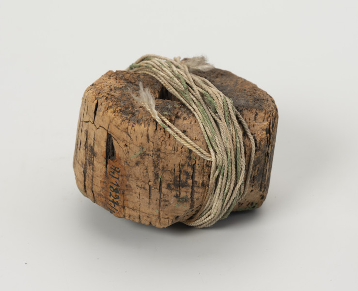 węda pływająca - Ujęcie z boku; Węda pływająca składająca się z okrągłego pływaka z otworem w środku, wykonanego z korka, na powierzchni którego widać liczne obłupienia. Do pływaka przywiązana jest bawełniana nić.