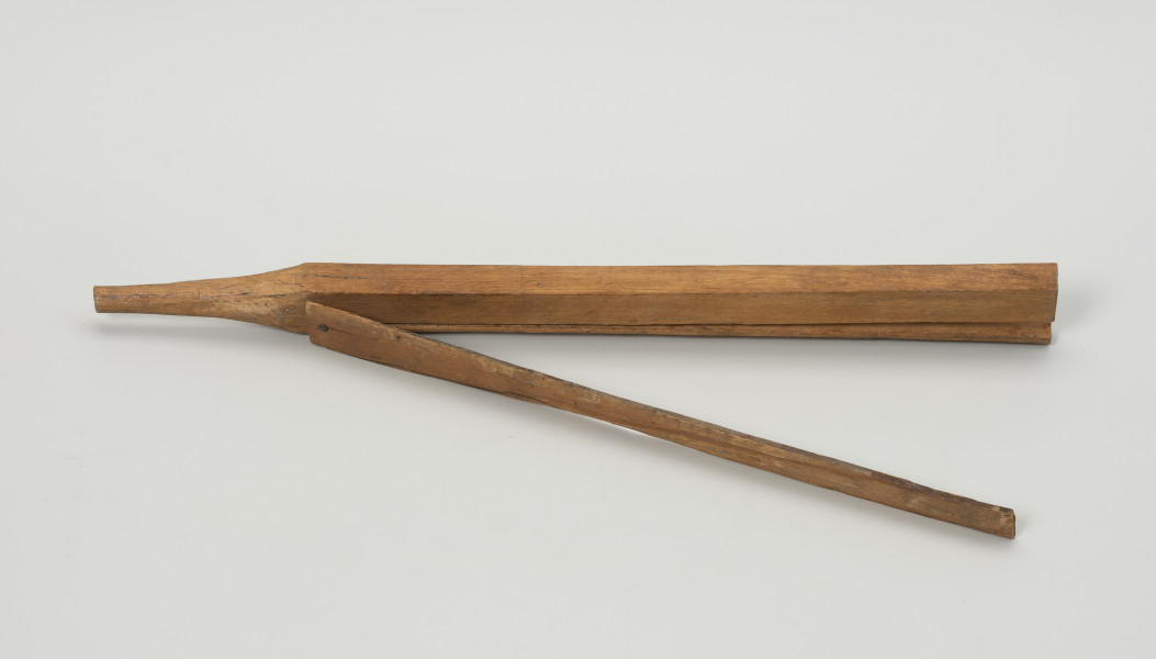 klamra do haczyków - Ujęcie z boku; Drewniana klamra ma kształt podłużnej listewki o czworobocznym przekroju. jeden z końców rozszczepiono do 3/4 długości. Drugi z lewej zaostrzono na końcu nadając mu stożkowaty kształt pasujący do otworu w ramieniu stojaka, w które wkładano sznurki z zawieszonymi na nich zestawami haczyków do suszenia. Nad szczeliną umieszczona jest, mocowana na gwoździk, płaska listewka dociskowa.