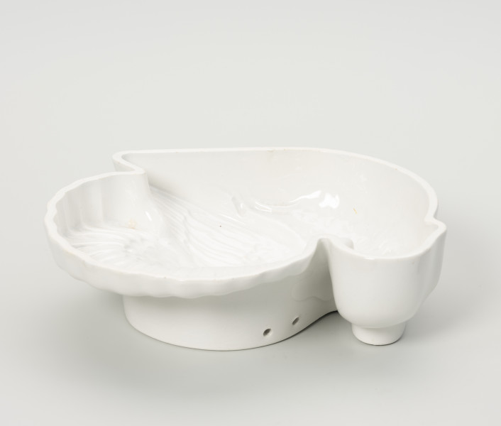 forma ceramiczna - Ujęcie z tyłu; Fajansowe, szkliwione jednolicie na biało naczynie w kształcie indyka. Realistyczna postać ptaka odwzorowana w postaci wgłębnego wzoru. Dno z wyraźnie wyodrębnioną stopką w części środkowej. Na stopce dwa otwory dające możliwość powieszenia naczynia na ścianie lub w kredensie.
