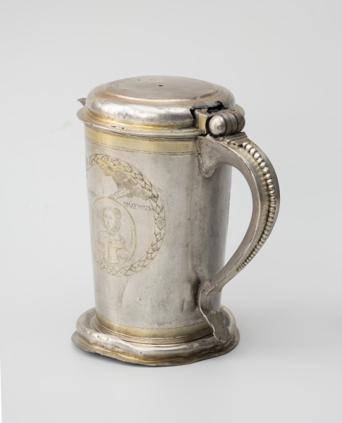 dzban na wino komunijne - Ujęcie tyłu ze skosu z lewej; Cylindryczny dzban o korpusie lekko zwężającym się ku dołowi, wsparty na wysklepionej stopie i nakryty spłaszczoną pokrywą na zawiasie, z utrąconym uchwytem. Esowato wygięte ucho ozdobione perełkowaniem i zakończone płaską tarczką.
Korpus dzbana zdobi grawerowana i złocona dekoracja. W wieńcu z liści laurowych medalion portretowy trzymany przez wysuwającą się z obłoków rękę Boga. W owalu młoda kobieta o włosach gładko zaczesanych nad czołem i ujętych w koczek, z drobnymi loczkami przy twarzy, w odsłaniającej ramiona sukni z bufiastymi rękawami, na jej szyi okrągły medalion i naszyjnik z pereł. Zgodnie z podpisem portret przedstawia Elisabethę Brüsewitz.