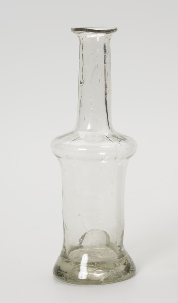 karafinka - Ujęcie z tyłu; Niewielka butelka o pojemności 0,25 L. Wydmuchana na piszczelu z bezbarwnego szkła – na brzuścu widoczne smugi z nie do końca prawidłowego procesu produkcji. Kształt podłużny, wąski, o grubym, wyraźnie wyodrębnionym dnie, brzusiec w górnej części uwypuklony przechodzi w wąską, długą szyjkę zakończoną płasko wywiniętym niewielkim wylewem. Na obwodzie podstawy z tyłu na tym ujęciu odciśnięty, owalny stempel.
Korka brak.