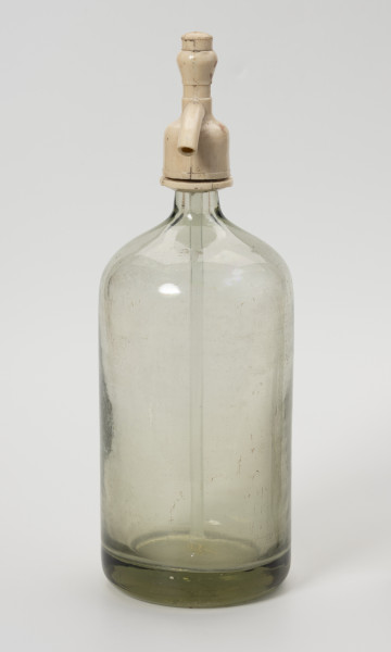 syfon do wody sodowej - Ujęcie z prawej; Grubościenna szklana butla o pogrubionym dnie i krótkiej, wąskiej szyjce z bezbarwnego szkła zwieńczona zamknięciem wykonanym z jasnego tworzywa sztucznego, składającego się z kraniku i ruchomej rączki niwidocznej na tym ujęciu połączonej ze szklaną rurką wpuszczoną do wnętrza butli. Po naciśnięciu rączki zamknięcia, leci woda gazowana.