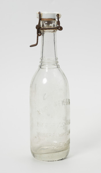 butelka 0,35 L z zamknięciem kabłąkowym - Ujęcie ze skosu z lewej; Butelka ze szkła niebarwionego, wytłaczany w formie. Szyjka prosta zakończona pogrubionym wlewem z kołnierzem. Na przejściu szyjki w brzusiec trzy wypukłe pierścienie. Brzusiec prosty przechodzi bezpośrednio w grube dno. Dno w części środkowej wklęsłe. W kołnierzu szyjki osadzone druciane zamknięcie z ceramicznym korkiem z gumową uszczelką. Na brzuścu wypukły napis: 