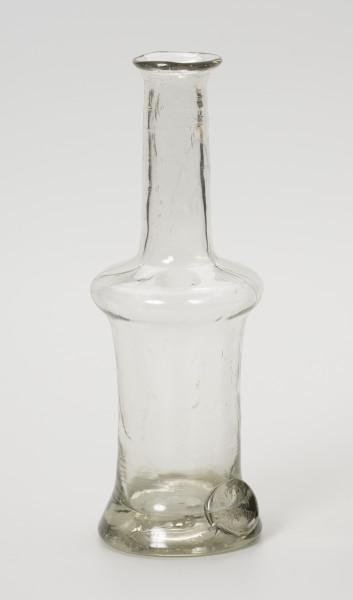 karafinka - Ujęcie ze skosu z lewej; Niewielka butelka o pojemności 0,25 L. Wydmuchana na piszczelu z bezbarwnego szkła – na brzuścu widoczne smugi z nie do końca prawidłowego procesu produkcji. Kształt podłużny, wąski, o grubym, wyraźnie wyodrębnionym dnie, brzusiec w górnej części uwypuklony przechodzi w wąską, długą szyjkę zakończoną płasko wywiniętym niewielkim wylewem. Na obwodzie podstawy w formie odciśniętego, owalnego stempla umieszczony napis: 