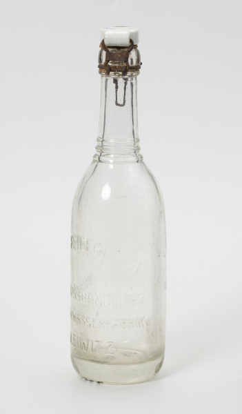 butelka 0,35 L z zamknięciem kabłąkowym - Ujęcie ze skosu z prawej; Butelka ze szkła niebarwionego, wytłaczany w formie. Szyjka prosta zakończona pogrubionym wlewem z kołnierzem. Na przejściu szyjki w brzusiec trzy wypukłe pierścienie. Brzusiec prosty przechodzi bezpośrednio w grube dno. Dno w części środkowej wklęsłe. W kołnierzu szyjki osadzone druciane zamknięcie z ceramicznym korkiem z gumową uszczelką. Na brzuścu wypukły napis.