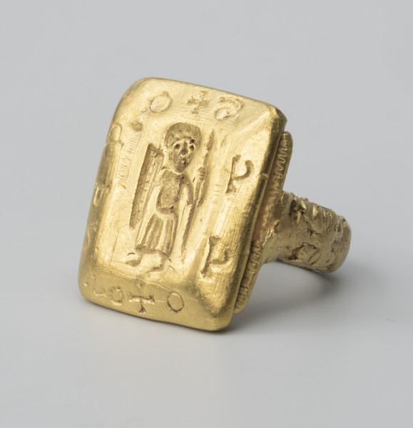 pierścień z Pęzina - Ujęcie ze skosu z prawej; Pierścień zbudowany z obrączki i dużej prostokątnej tarczki. Zewnętrzna część obrączki, lekko wypukła, ukształtowana została w formie dwóch antytetycznie ułożonych lwów dotykających paszczami tarczki. Tarczka prostokątna, zbliżona do kwadratu, o fasetowanych bokach. Wzdłuż boków łączących się z obrączką, wąskie strzemiączka ozdobione nacinaniem. W środkowej części tarczki, we wklęsłym reliefie, postać stojącego mężczyzny w długiej szacie, z włócznią i podłużną, trójkątną tarczą w dłoniach. Na sfazowanych brzegach tarczki nierozpoznana inskrypcja, składająca się z 10 znaków rozdzielonych dwoma znakami krzyża greckiego.