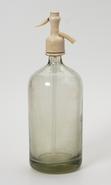 syfon do wody sodowej - Ujęcie z przodu; Grubościenna szklana butla o pogrubionym dnie i krótkiej, wąskiej szyjce z bezbarwnego szkła zwieńczona zamknięciem-pompką wykonanym z jasnego tworzywa sztucznego, składającego się z kraniku z prawej i ruchomej rączki z lewej połączonej ze szklaną rurką wpuszczoną do wnętrza butli. Po naciśnięciu rączki zamknięcia, leci woda gazowana.