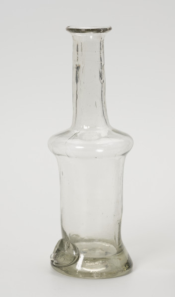 karafinka - Ujęcie ze skosu z prawej; Niewielka butelka o pojemności 0,25 L. Wydmuchana na piszczelu z bezbarwnego szkła – na brzuścu widoczne smugi z nie do końca prawidłowego procesu produkcji. Kształt podłużny, wąski, o grubym, wyraźnie wyodrębnionym dnie, brzusiec w górnej części uwypuklony przechodzi w wąską, długą szyjkę zakończoną płasko wywiniętym niewielkim wylewem. Na obwodzie podstawy w formie odciśniętego, owalnego stempla umieszczony napis: 