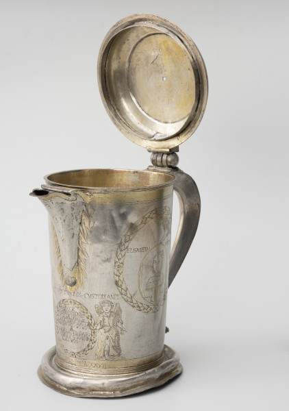 dzban na wino komunijne - Ujęcie ze skosu z prawej; Cylindryczny dzban o korpusie lekko zwężającym się ku dołowi, wsparty na wysklepionej stopie z pokrywą w tym ujeciu otwartą spłaszczoną na zawiasie. Dwa niewielkie otwory w pokrywie wskazują na pierwotne zwieńczenie, prawdopodobnie o charakterze figuralnym. Wylew trójkątny, faliście wycięty, częściowo przesłonięty. Esowato wygięte ucho ozdobione perełkowaniem i zakończone płaską tarczką.
Korpus dzbana zdobi grawerowana i złocona dekoracja. Na bokuw wieńcu z liści laurowych medalion portretowy. Poniżej wylewu lekko z prawej anioł trzymający wieniec laurowy z inskrypcją fundacyjną.