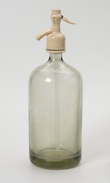 syfon do wody sodowej - Ujęcie z tyłu; Grubościenna szklana butla o pogrubionym dnie i krótkiej, wąskiej szyjce z bezbarwnego szkła zwieńczona zamknięciem-pompką wykonanym z jasnego tworzywa sztucznego, składającego się z kraniku z lewej i ruchomej rączki z prawej połączonej ze szklaną rurką wpuszczoną do wnętrza butli. Po naciśnięciu rączki zamknięcia, leci woda gazowana.