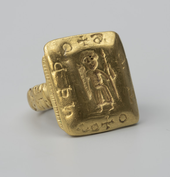 pierścień z Pęzina - Ujęcie ze skosu z lewej; Pierścień z dużą prostokątną tarczką. W środkowej części tarczki, we wklęsłym reliefie, postać stojącego mężczyzny w długiej szacie, z włócznią i podłużną, trójkątną tarczą w dłoniach. Na sfazowanych brzegach tarczki nierozpoznana inskrypcja, składająca się z 10 znaków rozdzielonych dwoma znakami krzyża greckiego.