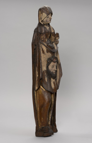 figura ołtarzowa; Święta Weronika - Ujęcie ze skosu z lewej; Smukła kobieca figura stojąca frontalnie o wydłużonej sylwetce i drobnej twarzy, polichromowana i złocona. Święta, ubrana w suknię z wysokim stanem, przewiązaną luźno opuszczonym pasem z klamrą oraz w długi płaszcz, trzyma przed sobą chustę z reliefowym wizerunkiem głowy Chrystusa w koronie cierniowej. Niewielką głowę figury otula chusta spływającą na ramiona i marszczona pod szyją podwiką. Twarz, o oczach zapatrzonych w dal i lekko pochylona w lewą stronę, wyraża smutek. Na prawym policzku świętej wydrążona rana cięta. Tył płaski, typowy dla rzeźb przyściennych. Brak prawej dłoni. Wyłamania drewna przy podstawie. Znaczne ubytki polichromii i złoceń.