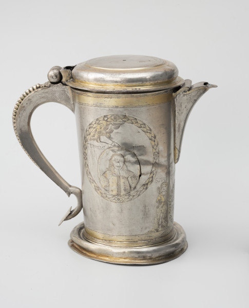 dzban na wino komunijne - Ujęcie z lewej; Cylindryczny dzban o korpusie lekko zwężającym się ku dołowi, wsparty na wysklepionej stopie i nakryty spłaszczoną pokrywą na zawiasie. Wylew trójkątny, faliście wycięty, częściowo przesłonięty. Esowato wygięte ucho ozdobione perełkowaniem i zakończone płaską tarczką. Korpus dzbana zdobi grawerowana i złocona dekoracja. W wieńcu z liści laurowych medalion portretowy trzymany przez wysuwającą się z obłoków rękę Boga, ukazany jest mężczyzna o długich, lekko falujących włosach, z niewielkim podkręconym wąsem oraz bródką, ubrany w wams zapinany na szereg guzików, z wyłożonym kołnierzem. Przy portrecie mężczyzny napis TEOPHILVS PIPER