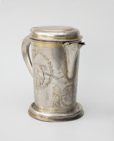 dzban na wino komunijne - Ujęcie ze skosu z lewej; Cylindryczny dzban o korpusie lekko zwężającym się ku dołowi, wsparty na wysklepionej stopie i nakryty spłaszczoną pokrywą. Wylew trójkątny, faliście wycięty, częściowo przesłonięty. Esowato wygięte ucho. Korpus dzbana zdobi grawerowana i złocona dekoracja. Z bok z lewej w wieńcu z liści laurowych medalion portretowy, mężczyzna o długich, lekko falujących włosach, z niewielkim podkręconym wąsem oraz bródką, ubrany w wams zapinany na szereg guzików, z wyłożonym kołnierzem. Wylew w otoczeniu płomieni, poniżej anioł trzymający wieniec laurowy z inskrypcją fundacyjną. SIMPLEX .ET. RECTVM NOS.CVSTODIANT | HOCCE. |  MONVMENTVM |  ECCLESIAE SPIRIT SANCTI.SIC. DICTAE | AD.SACROS. VSVS. |  A  | PRAEDICTO. PARI |  CONIV. GVMOB: |  LATVM | A.O.R.M DCLXXVII