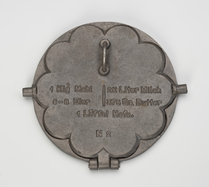 forma do wypieku gofrów - Ujęcie z góry; Forma do wypieku gofrów wykonana z żeliwa. Składa się z okrągłej podstawy oraz ściśle przylegającej do niej pokrywy w kształcie wielopłatkowego kwiatka. Na pokrywie napis w języku niemieckim (przepis na ciasto oraz oznaczenie literowo-liczbowe). Nad napisem zamontowany półkolisty uchwyt. Zewnętrzna powierzchnia podstawy gładka. Żeliwna obręcz umożliwiająca położenie formy na fajerkach nie zachowała się.