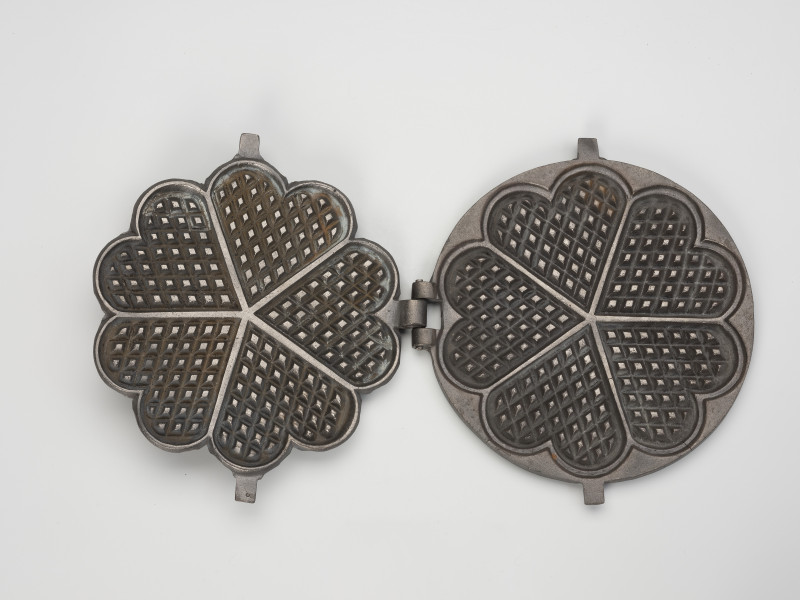 forma do wypieku gofrów - Ujęcie środka; Forma do wypieku gofrów wykonana z żeliwa. Składa się z okrągłej podstawy oraz ściśle przylegającej do niej pokrywy w kształcie wielopłatkowego kwiatka. Obie powierzchnie pokryte wewnątrz kratownicą, podzieloną na 5 jednakowych pół w kształcie serca. Żeliwna obręcz umożliwiająca położenie formy na fajerkach nie zachowała się.