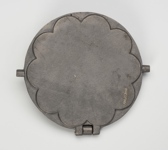 forma do wypieku gofrów - Ujęcie z dołu; Forma do wypieku gofrów wykonana z żeliwa. Składa się z okrągłej podstawy oraz ściśle przylegającej do niej pokrywy w kształcie wielopłatkowego kwiatka. Zewnętrzna powierzchnia podstawy gładka. Żeliwna obręcz umożliwiająca położenie formy na fajerkach nie zachowała się.