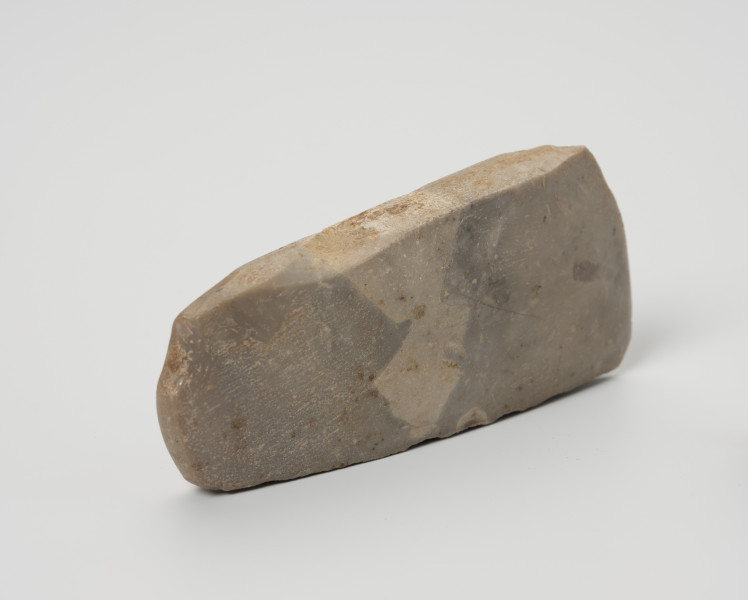 siekiera krzemienna - Ujęcie ze skosu; Siekiera czworościenna o grubym obuchu i lekko skośnym ostrzu, szlifowana i gładzona na całej powierzchni, wykonana z krzemienia kredowego