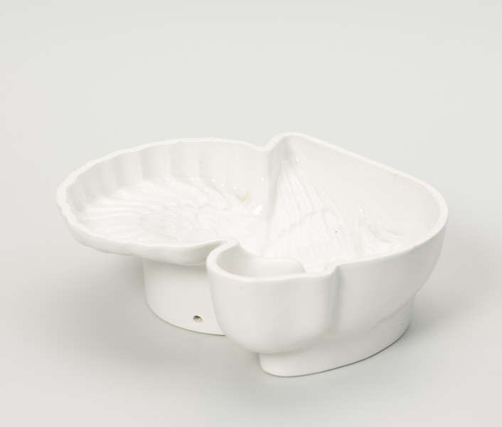 tłok pieczętny - Ujęcie z boku; Fajansowe, szkliwione jednolicie na biało naczynie w kształcie indyka. Realistyczna postać ptaka odwzorowana w postaci wgłębnego wzoru. Dno z wyraźnie wyodrębnioną stopką w części środkowej. Na stopce dwa otwory (na ujęciu widoczny tylko jeden) dające możliwość powieszenia naczynia na ścianie lub w kredensie.