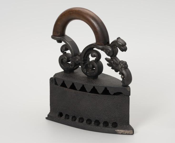 żelazko na węgiel - Ujęcie przodu z skosu z lewej; Żelazko na węgiel wykonane z żeliwa (odlew). Stopa o kształcie przypominającym trójkąt równoramienny z lekko zaokrąglonymi dłuższymi bokami. Korpus – pojemnik na węgiel – wysoki o prostych ściankach. Wzdłuż jego dolnych, dłuższych krawędzi niewielkie, okrągłe otwory. Klapka przykręcona do korpusu za pomocą śruby. Pokrywa uchylna, mocowana na znajdującym się z tyłu korpusu metalowym pręcie. Do zewnętrznej strony mocowany za pomocą dwóch śrub ozdobny uchwyt. Przednia część uchwytu zdobiona głową smoka jest ruchoma, z rozwidlonym końcem, zaczepianym o mocowany w korpusie pręcik zakończony gałką.