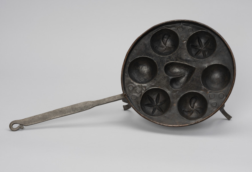 patelnia dołkownica - Ujęcie z góry; Patelnia z miedzianym naczyniem oraz kutymi z żelaza: rączką i nóżkami. Talerz patelni okrągły, miedziany o niskim kołnierzu i z siedmioma wgłębieniami na ciasto. Środkowe wgłębienie w kształcie serca, pozostałe, okalające je – okrągłe. W dwóch z nich wzór kwiatowy, w dwóch po trzy, schematycznie przedstawione ptaszki (obecnie wzór częściowo zatarty). Nóżki dołączone od spodu wykonane z płaskowników, u dołu esowato wygięte na zewnątrz. Do jednej z nóżek mocowana długa, prosta rączka. Na około 2/3 długości spłaszczona. Koniec rączki wygięty esowato i dociśnięty do uchwytu, tworząc otwór w kształcie łezki. Patelnia nosi ślady użytkowania.