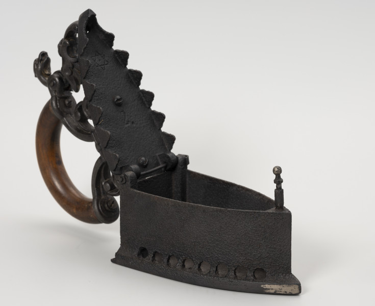 żelazko na węgiel - Ujęcie z przodu z uchylną klapą; Żelazko na węgiel wykonane z żeliwa (odlew). Stopa o kształcie przypominającym trójkąt równoramienny z lekko zaokrąglonymi dłuższymi bokami. Korpus – pojemnik na węgiel – wysoki o prostych ściankach. Wzdłuż jego dolnych, dłuższych krawędzi niewielkie, okrągłe otwory. Klapka przykręcona do korpusu za pomocą śruby. Pokrywa uchylna, mocowana na znajdującym się z tyłu korpusu metalowym pręcie. Na wewnętrznej stronie pokrywy cyfra 2 oraz sześcioramienna, niewielka gwiazdka. Do zewnętrznej strony mocowany za pomocą dwóch śrub ozdobny uchwyt. Przednia część uchwytu zdobiona głową smoka jest ruchoma, z rozwidlonym końcem, zaczepianym o mocowany w korpusie pręcik zakończony gałką.