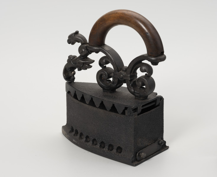 żelazko na węgiel - Ujęcie tyłu ze skosu z lewej; Żelazko na węgiel wykonane z żeliwa (odlew). Stopa o kształcie przypominającym trójkąt równoramienny z lekko zaokrąglonymi dłuższymi bokami. Korpus – pojemnik na węgiel – wysoki o prostych ściankach. Wzdłuż jego dolnych, dłuższych krawędzi niewielkie, okrągłe otwory. W ściance tylnej prostokątny, wąski otwór zamykany na ruchomą klapkę z niewielką gałką. Klapka przykręcona do korpusu za pomocą śruby. Pokrywa uchylna, mocowana na znajdującym się z tyłu korpusu metalowym pręcie. Do zewnętrznej strony mocowany za pomocą dwóch śrub ozdobny uchwyt. Przednia część uchwytu zdobiona głową smoka jest ruchoma, z rozwidlonym końcem, zaczepianym o mocowany w korpusie pręcik zakończony gałką.