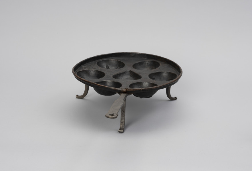 patelnia dołkownica - Ujęcie z tyłu; Patelnia z miedzianym naczyniem oraz kutymi z żelaza: rączką i trzema nóżkami. Talerz patelni okrągły, miedziany o niskim kołnierzu i z siedmioma wgłębieniami na ciasto. Środkowe wgłębienie w kształcie serca, pozostałe, okalające je – okrągłe. Trzy nóżki dołączone od spodu wykonane z płaskowników, u dołu esowato wygięte na zewnątrz. Do jednej z nóżek mocowana długa, prosta rączka. Na około 2/3 długości spłaszczona. Koniec rączki wygięty esowato i dociśnięty do uchwytu, tworząc otwór w kształcie łezki. Patelnia nosi ślady użytkowania.