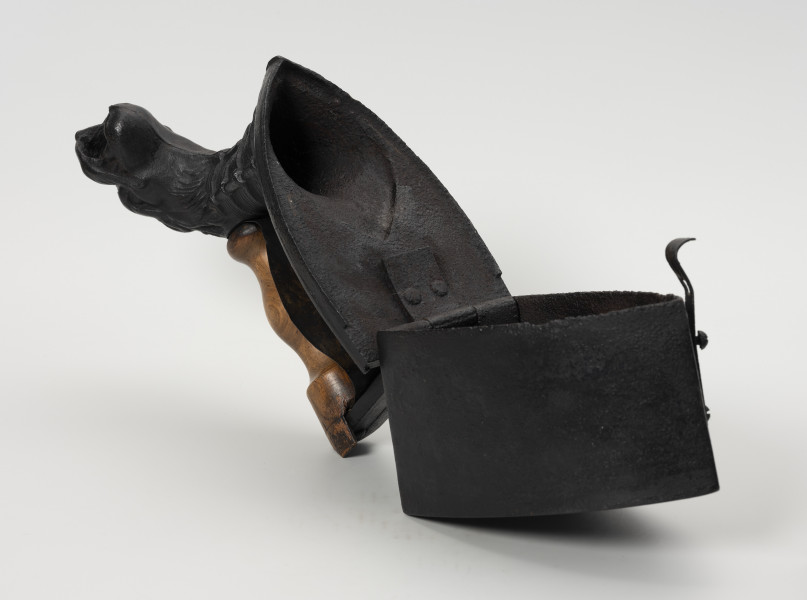 żelazko na węgiel z kominem - Ujęcie z przodu; Żelazko na węgiel z kominem wykonane z żeliwa (odlew). Pełniący rolę pojemnika na węgiel korpus żelazka wysoki, o prostych ściankach. Do tylnej ścianki korpusu mocowana za pomocą zawiasów uchylna pokrywa. W jej przedniej części znajduje się komin w kształcie głowy smoka z otwartą paszczą, w tylnej zaokrąglony u góry wspornik. Pomiędzy nimi zamontowana wykonana z drewna, profilowana rączka (częściowo uszkodzona).