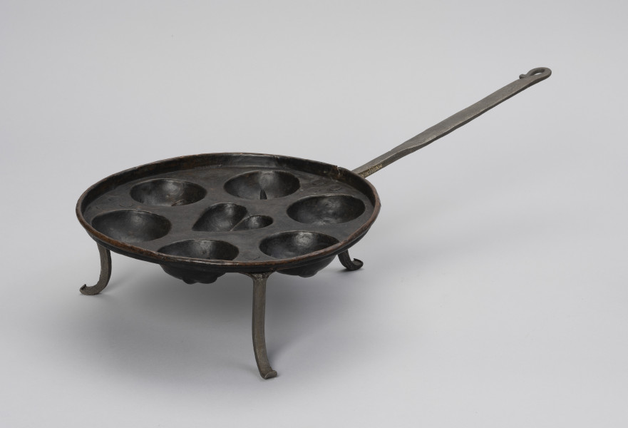 patelnia dołkownica - Ujęcie ze skosu z prawej; Patelnia z miedzianym naczyniem oraz kutymi z żelaza: rączką i trzema nóżkami. Talerz patelni okrągły, miedziany o niskim kołnierzu i z siedmioma wgłębieniami na ciasto. Środkowe wgłębienie w kształcie serca, pozostałe, okalające je – okrągłe. Trzy nóżki dołączone od spodu wykonane z płaskowników, u dołu esowato wygięte na zewnątrz. Do jednej z nóżek mocowana długa, prosta rączka. Na około 2/3 długości spłaszczona. Koniec rączki wygięty esowato i dociśnięty do uchwytu, tworząc otwór w kształcie łezki. Patelnia nosi ślady użytkowania.