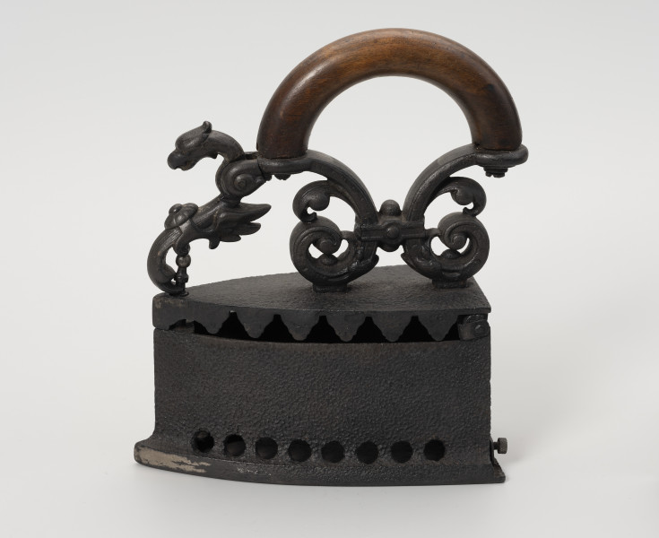 żelazko na węgiel - Ujęcie z drugiego boku; Żelazko na węgiel wykonane z żeliwa (odlew). Stopa o kształcie przypominającym trójkąt równoramienny z lekko zaokrąglonymi dłuższymi bokami. Korpus – pojemnik na węgiel – wysoki o prostych ściankach. Wzdłuż jego dolnych, dłuższych krawędzi niewielkie, okrągłe otwory. Klapka przykręcona do korpusu za pomocą śruby. Pokrywa uchylna, mocowana na znajdującym się z tyłu korpusu metalowym pręcie. Do zewnętrznej strony mocowany za pomocą dwóch śrub ozdobny uchwyt. Przednia część uchwytu zdobiona głową smoka jest ruchoma, z rozwidlonym końcem, zaczepianym o mocowany w korpusie pręcik zakończony gałką.