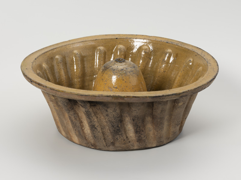 forma do wypieku babki - Ujęcie z przodu; Forma do wypieku o charakterystycznym dla wypieku babek kształcie. Brzusiec nieznacznie rozszerzający się ku górze, zdobiony lekko skośnymi, regularnymi karbami. Brzeg prosty, uformowany prostopadle do ścianek brzuśca. W centralnej części dna znajduje się komin o walcowatym kształcie, zakończony kopulasto z małym otworem na szczycie o zaznaczonym wylewie. Naczynie wykonane z gliny o jasnej barwie, szkliwo w jasnobrązowej tonacji pokrywa tylko jego wnętrze.